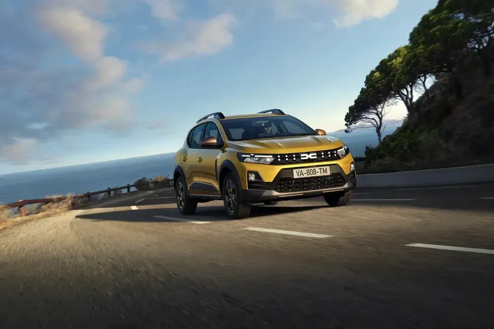 Dacia Sandero Stepway jaune sur route côtière, sous un ciel ensoleillé et bordé d'arbres.