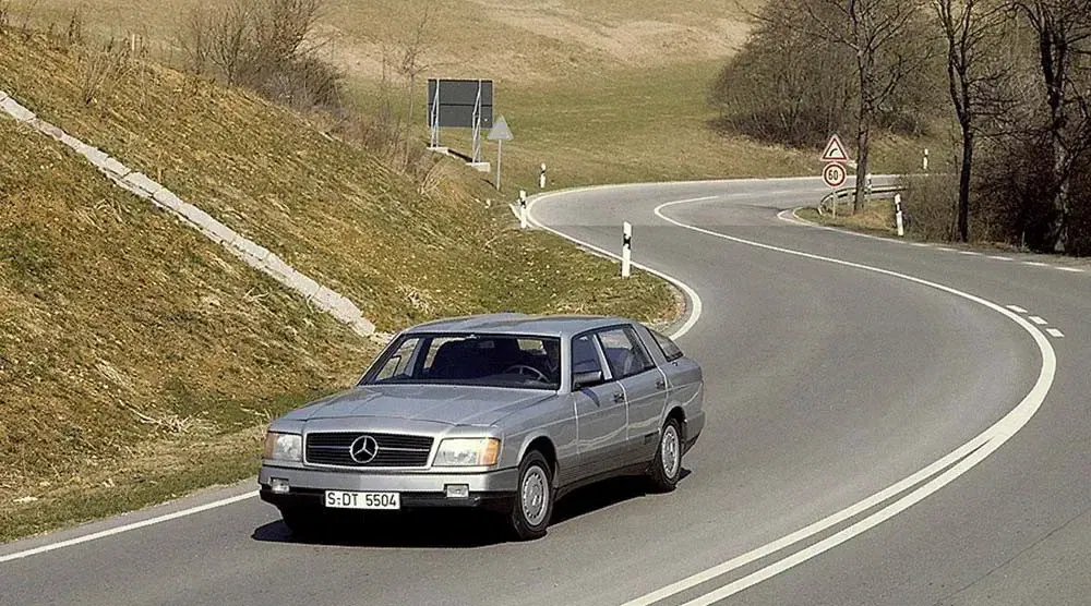 Klassieke zilveren Mercedes-Benz op kronkelige landweg met kale bomen op de achtergrond.