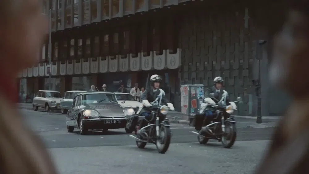 Citroën DS noire et motos de police dans une rue animée en milieu urbain.