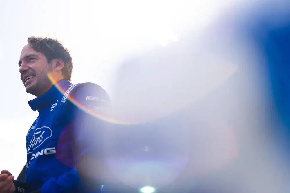 Pilote en combinaison Ford Racing souriant, éclairé par le soleil, légèrement flou.