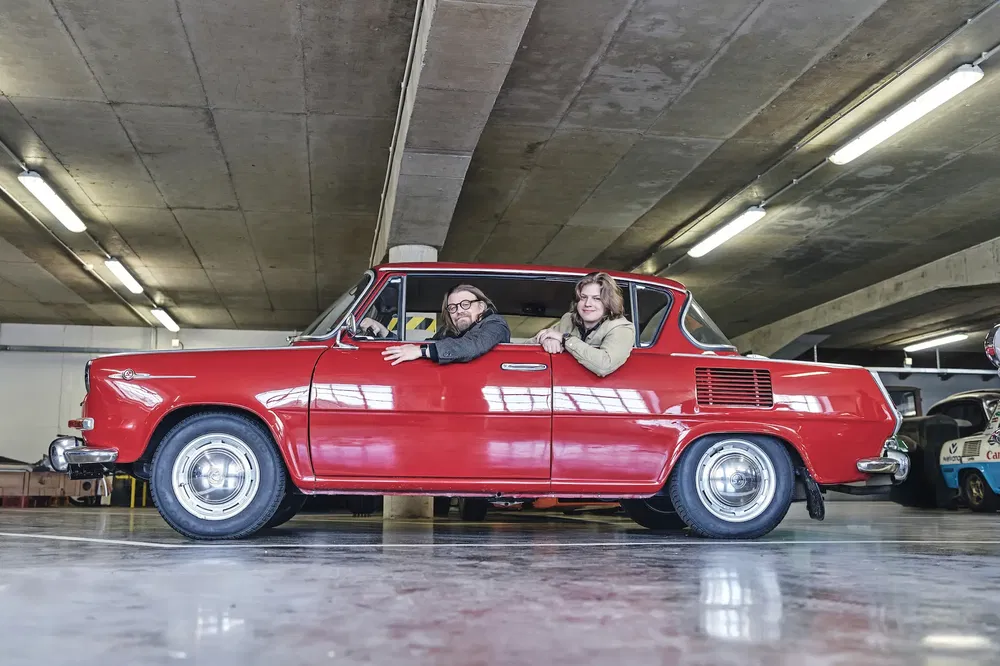 Skoda 1000 MB rouge dans un garage, deux personnes accoudées aux fenêtres.