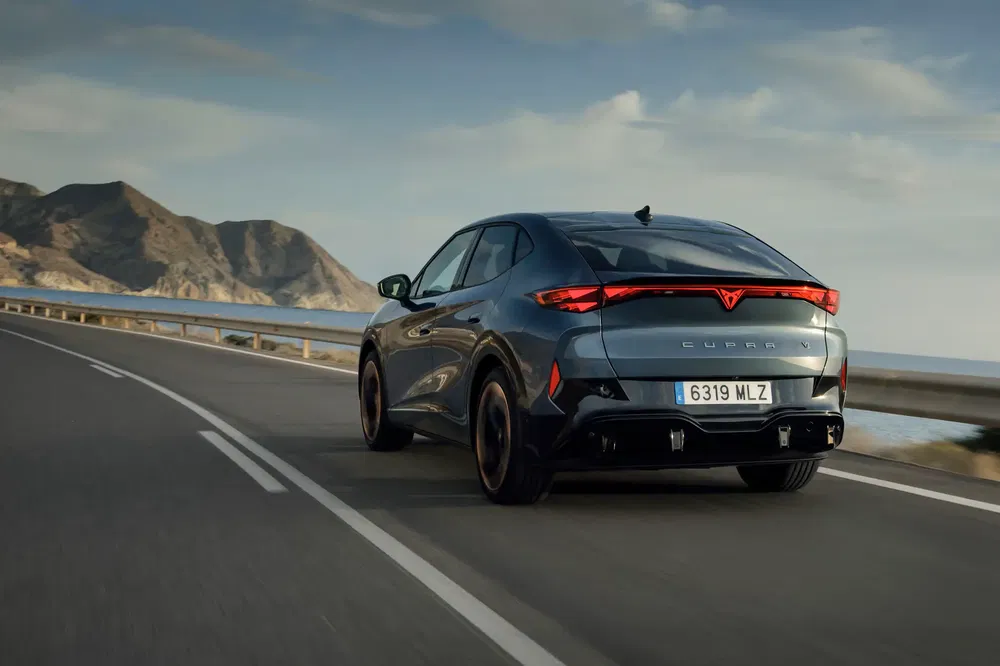 Zwarte Cupra auto rijdt op een kustweg met bergen op de achtergrond.