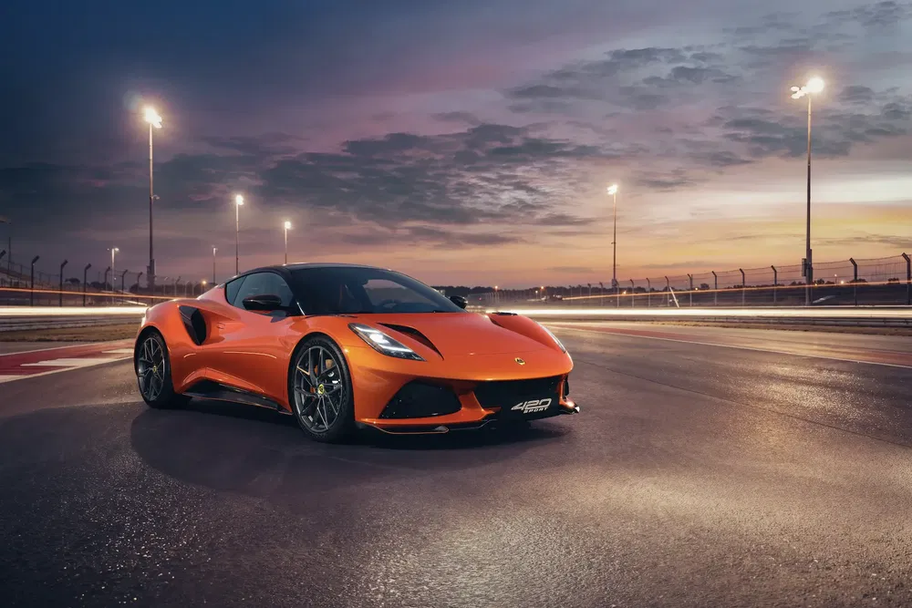 {"fr":"Voiture sportive Lotus Emira orange sur circuit au crépuscule, phares allumés.","nl":"Oranje Lotus Emira sportwagen op circuit bij zonsondergang, met lichten aan."}
