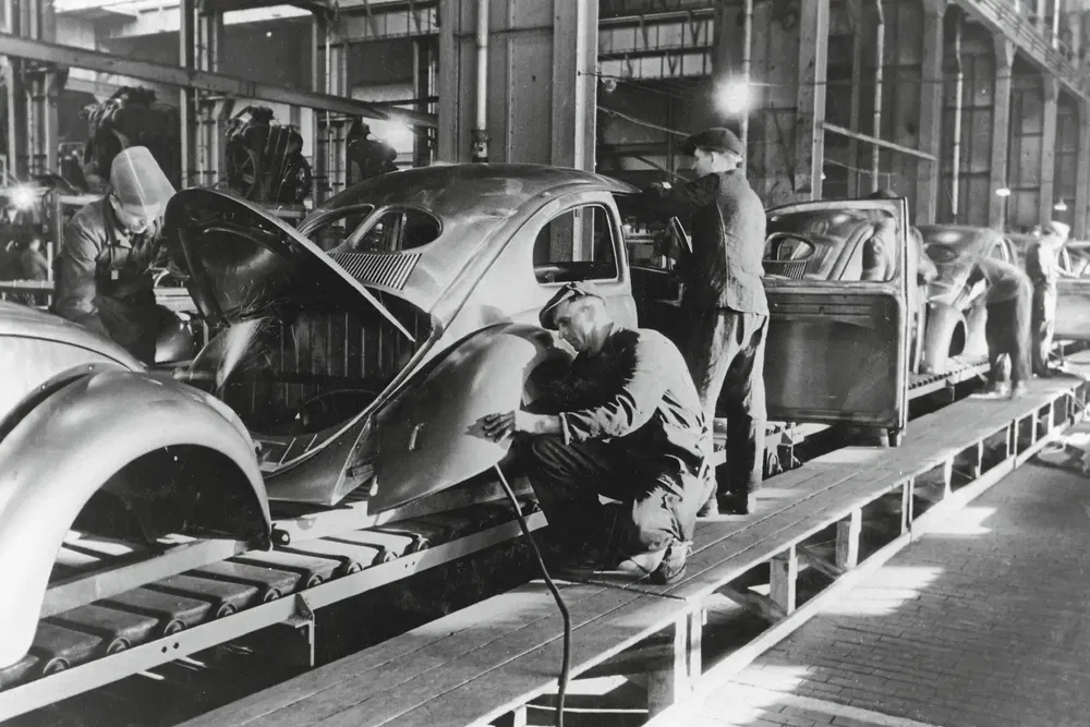 Chaîne de montage Volkswagen Beetle historique en usine, ouvriers assemblant carrosseries automobiles.