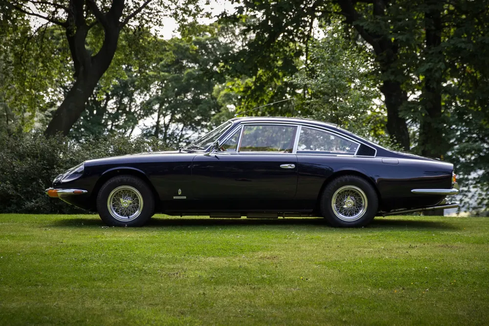 Zwarte Ferrari 365 GT 2+2 geparkeerd op groen grasveld, omgeven door loofbomen.