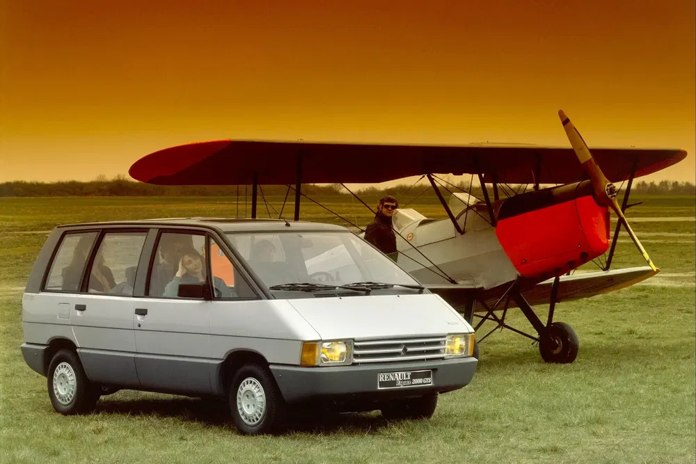 Renault Espace argentée devant un vieil avion biplan, ciel orangé crépusculaire.