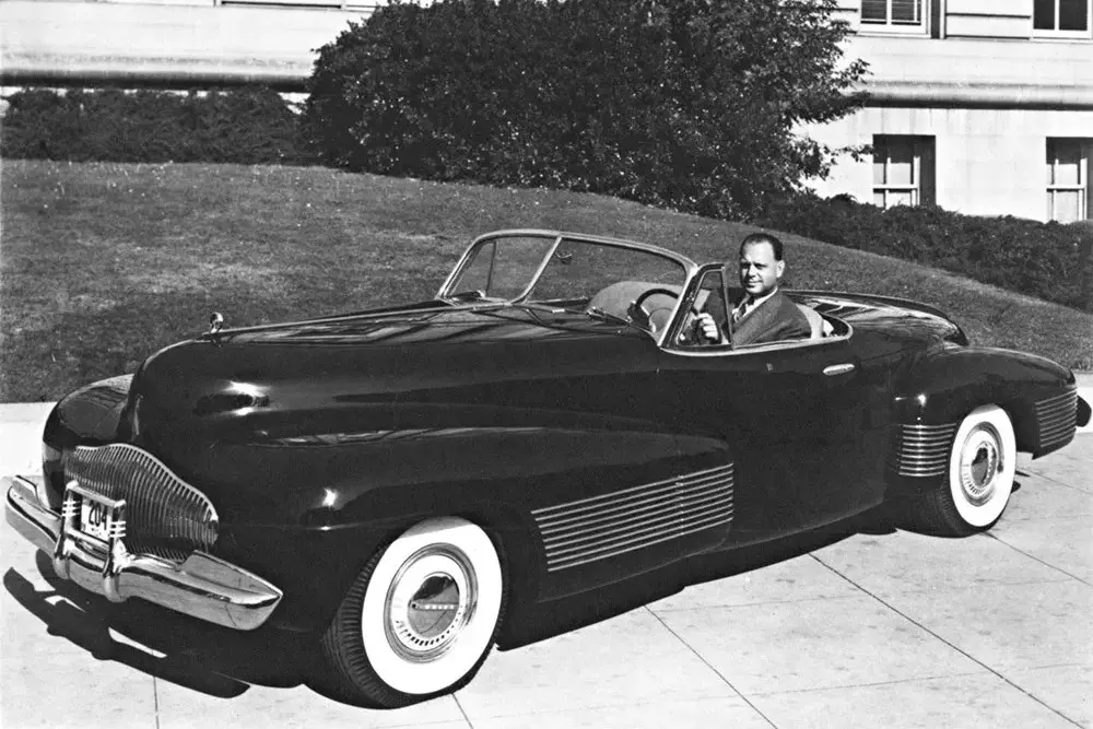 Buick convertible vintage avec pneus à flancs blancs, homme au volant sur une allée.