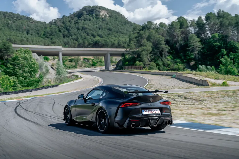 Zwarte Toyota Supra op kronkelweg omgeven door beboste heuvels.