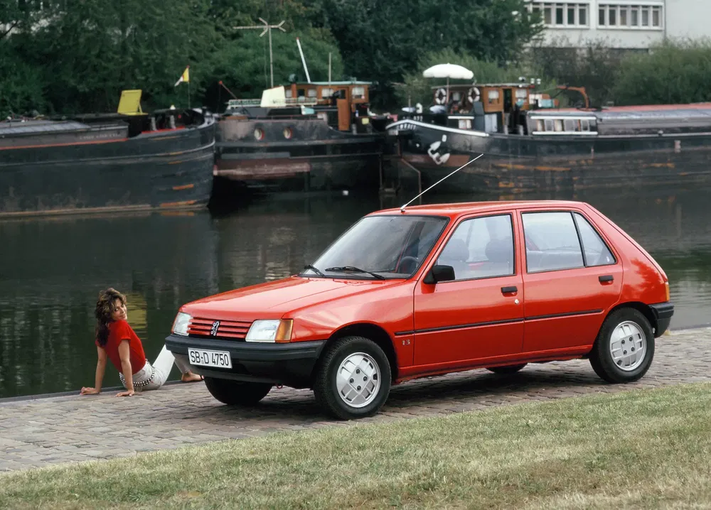 Peugeot 205 rouge près d'un canal, bateaux en arrière-plan, scène urbaine tranquille.