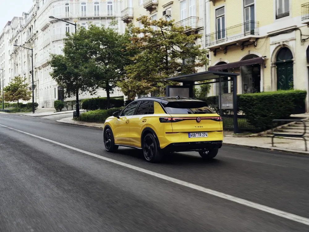 Gele Volkswagen T-Roc rijdt in stadsstraat met stijlvolle gebouwen aan weerszijden.