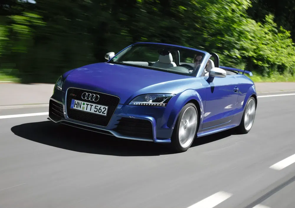 Audi TT RS cabriolet bleu roulant sur route bordée d'arbres.