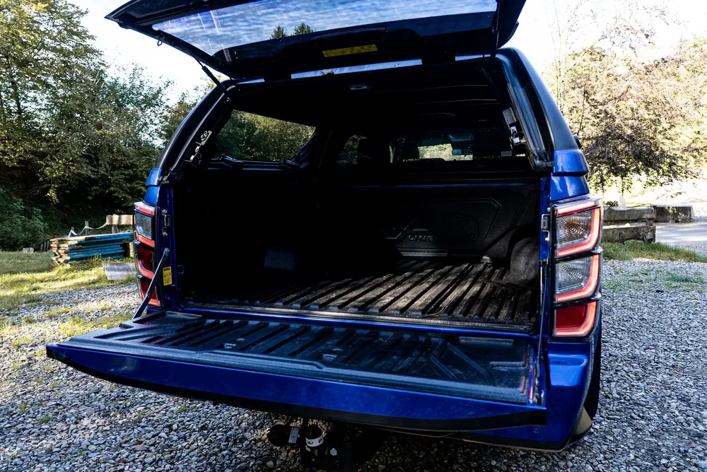 Coffre ouvert d'un pick-up bleu garé sur du gravier, avec espace de chargement spacieux.