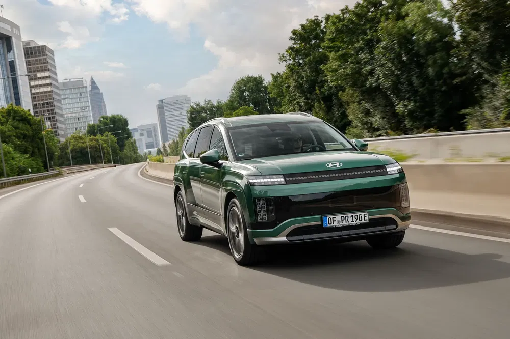 Hyundai Ioniq 7 vert roulant sur une autoroute urbaine bordée d'arbres et de bâtiments modernes.