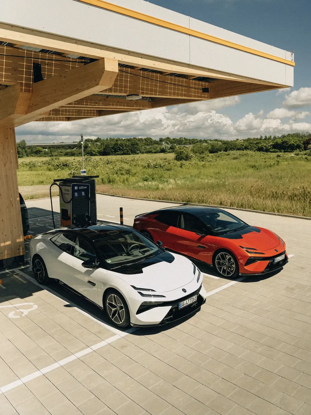 Deux voitures sportives électriques Lotus blanches et rouges stationnées à une borne de recharge rapide.