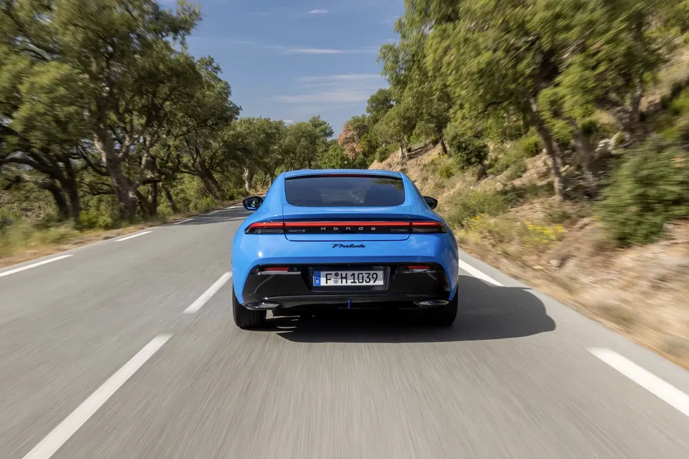 Achteraanzicht van een blauwe Porsche Taycan op een zonnige weg omgeven door bomen.