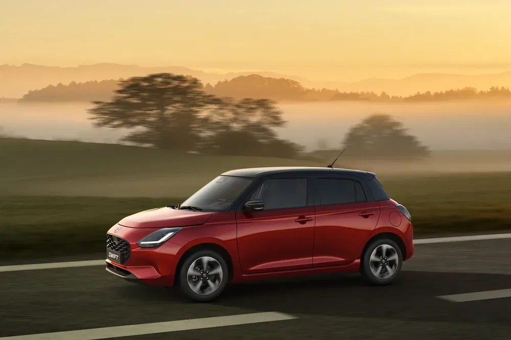 Nieuwe rode Suzuki Swift rijdt op weg, plattelandslandschap bij zonsopgang.