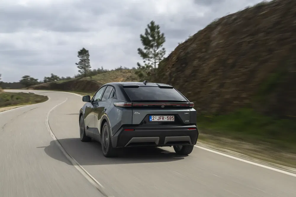 Grijze Toyota bZ4X rijdt op een kronkelige weg onder een bewolkte hemel.