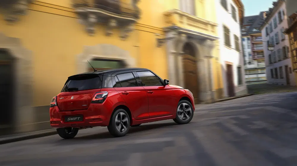 Rode Suzuki Swift rijdt door een schilderachtige straat met historische gebouwen.