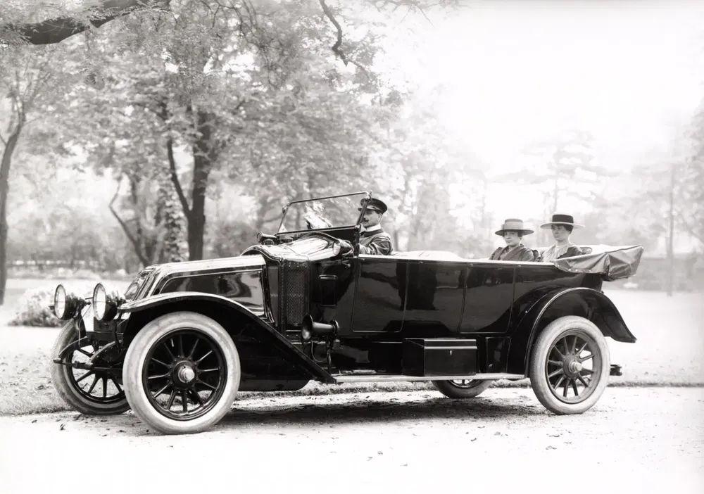Voiture ancienne décapotable avec passagers, dans un parc arboré en arrière-plan.