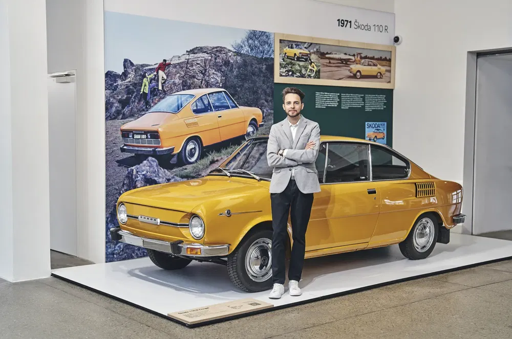 {"fr":"Skoda 110 R jaune exposée avec une affiche historique en arrière-plan dans un musée.","nl":"Gele Skoda 110 R tentoongesteld met historische poster op de achtergrond in een museum."}