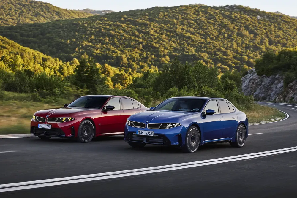 Deux BMW i5 électriques, rouge et bleue, sur une route sinueuse entourée de collines verdoyantes.