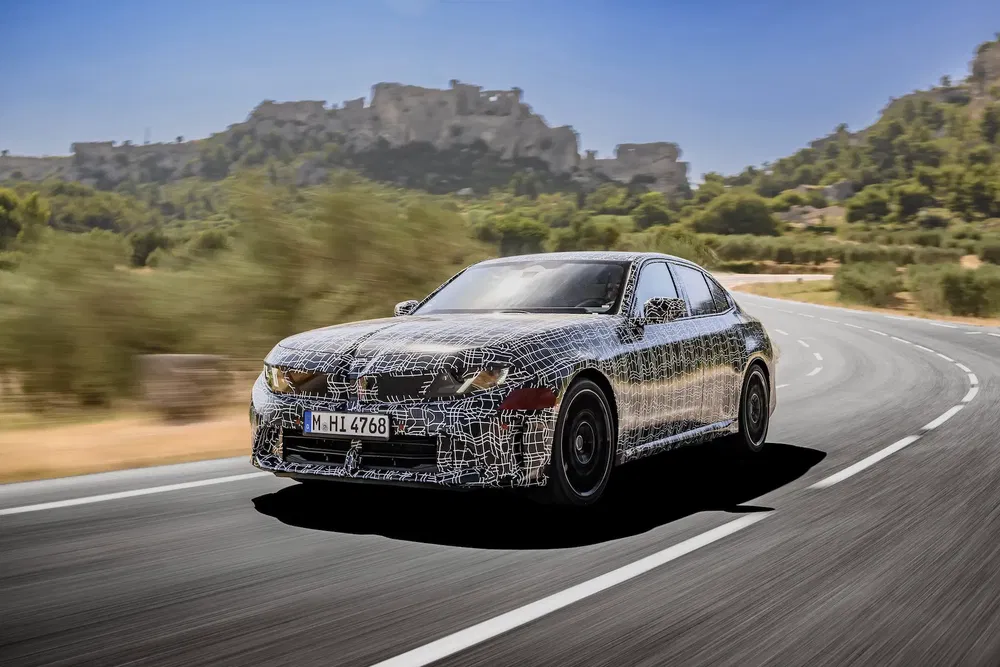 BMW camouflé en test routier, paysage montagneux en arrière-plan, conduite dynamique.