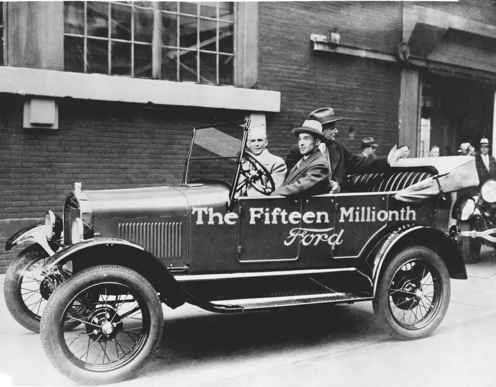 Ford Model T, quinzième millionième, avec passagers souriants devant une usine en brique.
