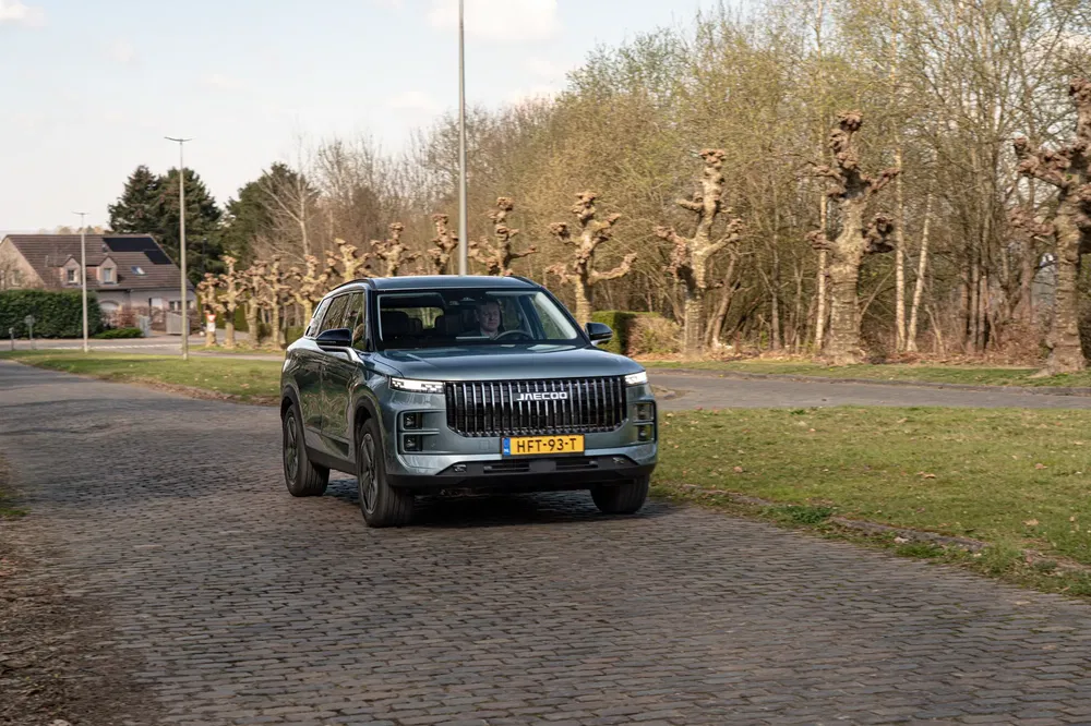 Moderne SUV rijdt op kinderkopjes met kale bomen op de achtergrond in landelijke omgeving.