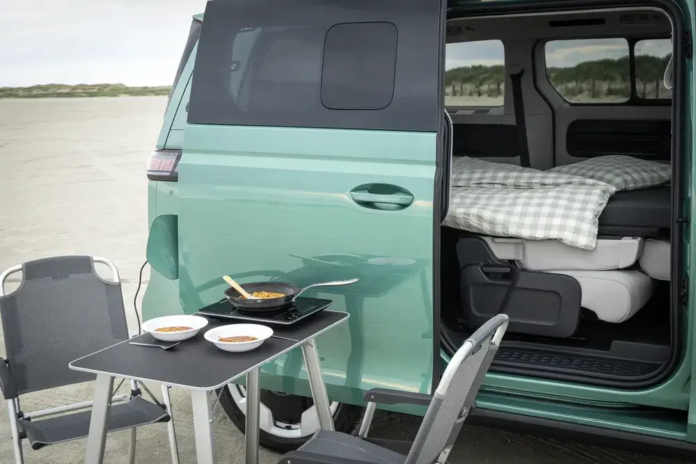 Van Volkswagen aménagé en camping, table extérieure avec chaises et repas préparé sur réchaud.