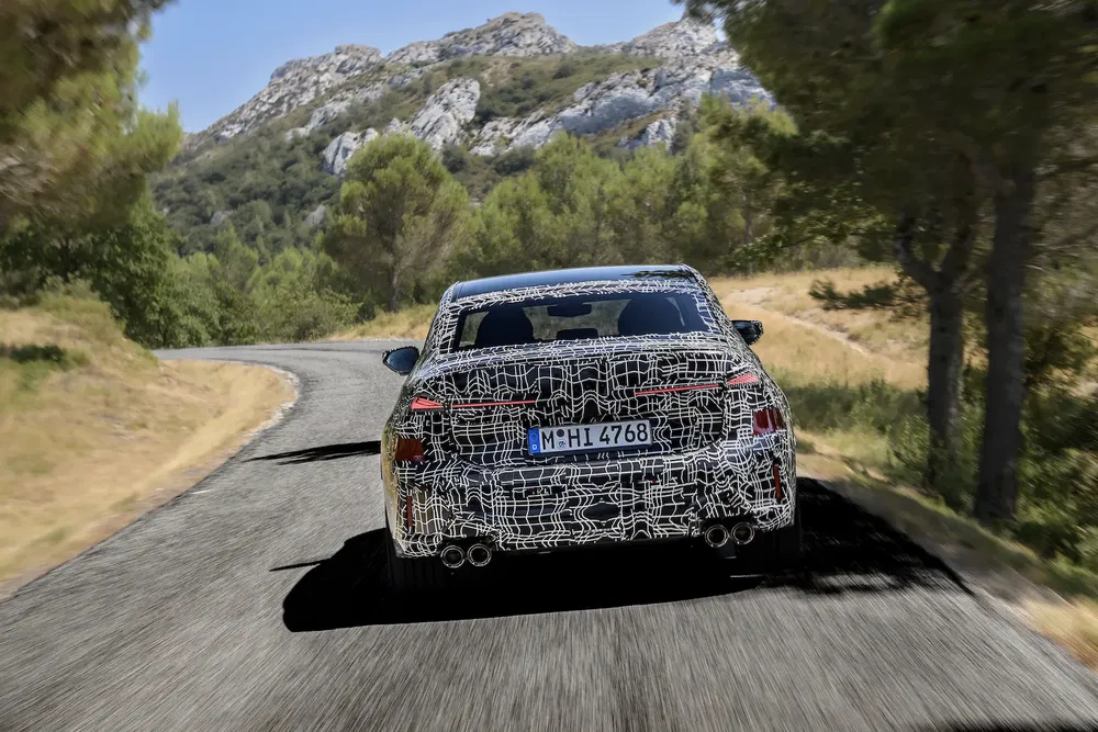 Prototype BMW camouflé sur route sinueuse entourée de forêt et montagnes.