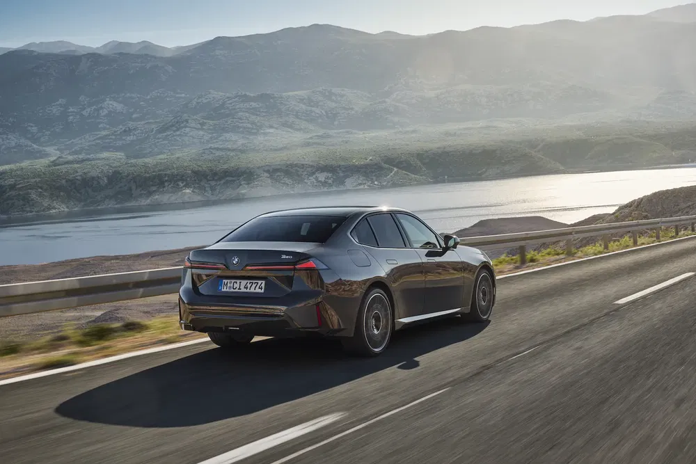BMW Série 3 roulant sur route côtière ensoleillée avec montagnes en arrière-plan.