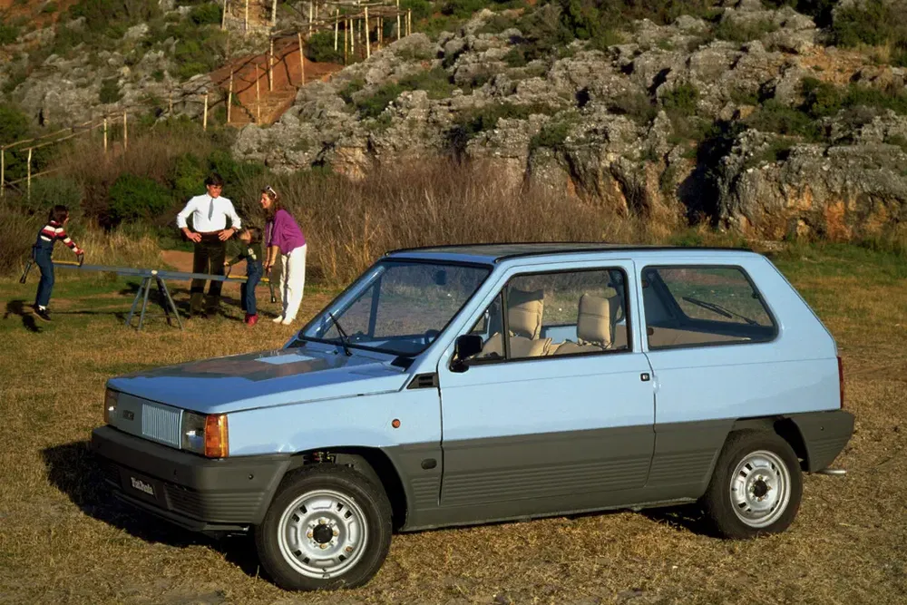 Vintage Fiat Panda voor rotsachtige heuvel, spelende familie op de achtergrond.