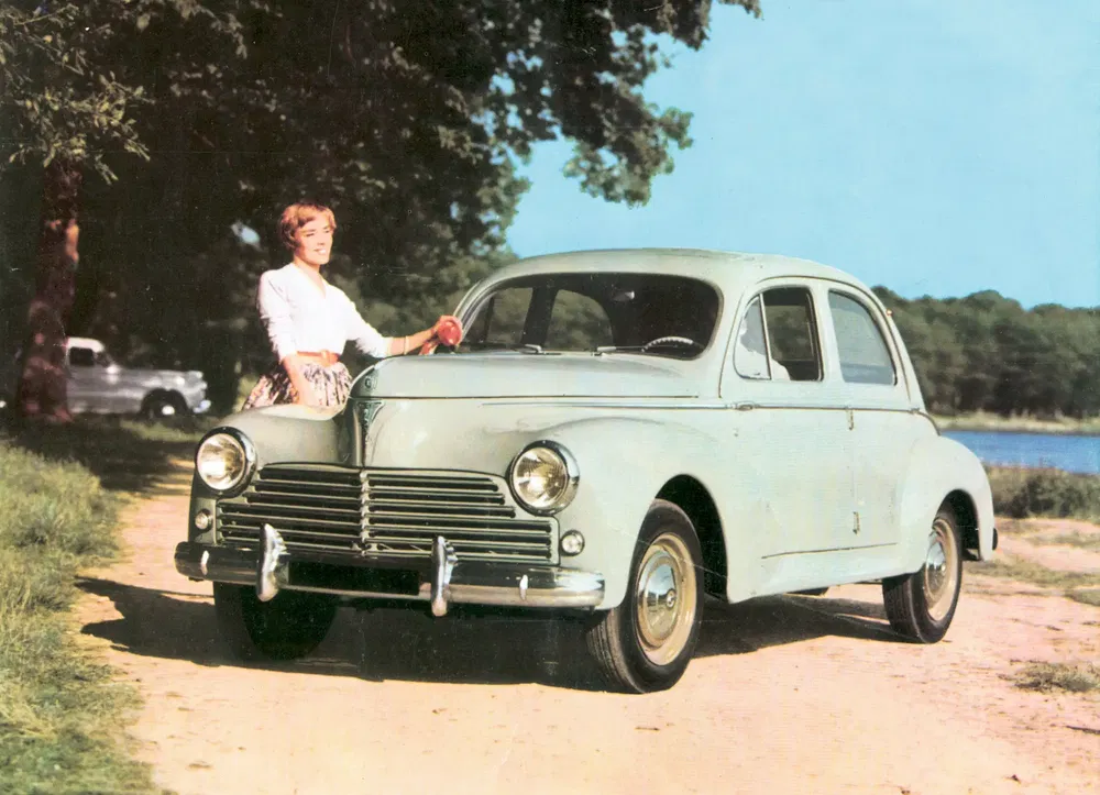 Voiture Peugeot 203 vintage verte clair avec femme souriante, nature en arrière-plan, ciel bleu.
