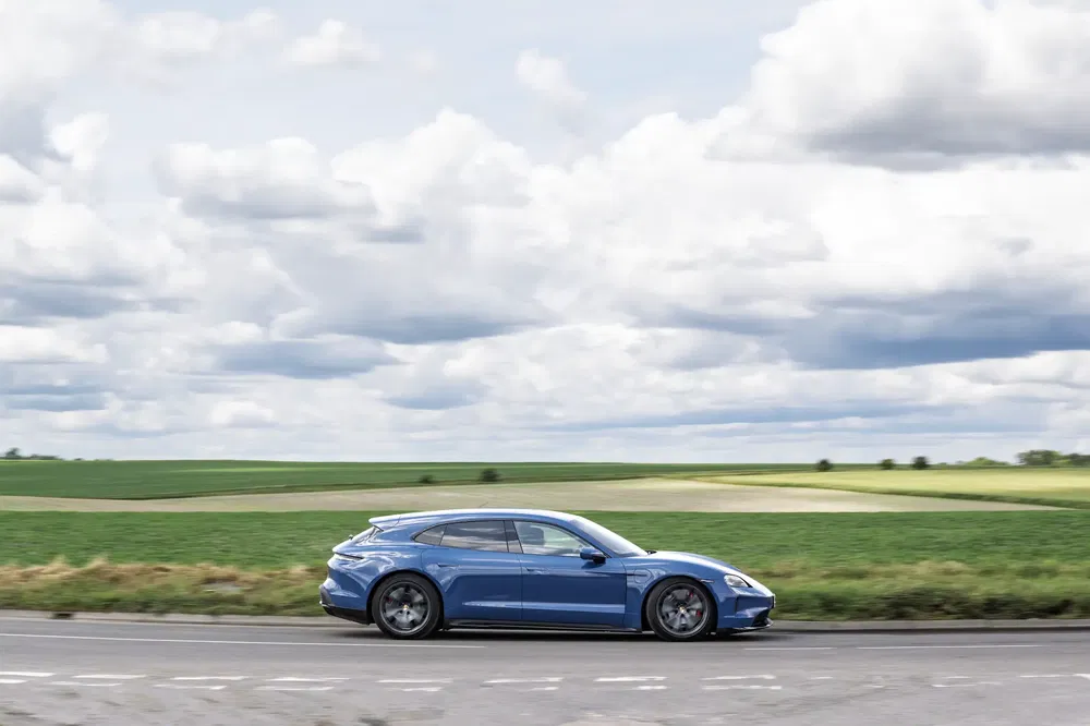 Voiture Porsche bleue roulant sur une route de campagne sous ciel nuageux.