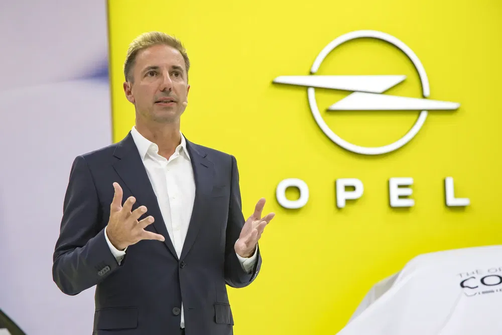 Man in pak spreekt voor geel Opel-logo tijdens een autoconferentie.