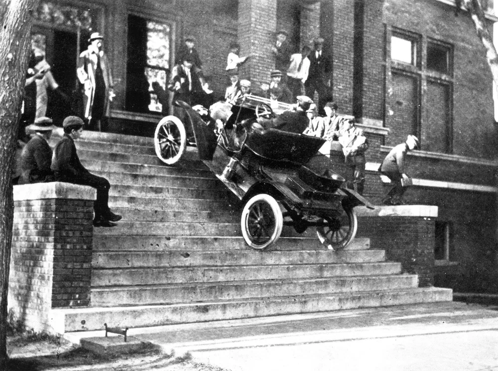 Voiture ancienne descendant des escaliers, entourée de spectateurs curieux sur un bâtiment en briques.
