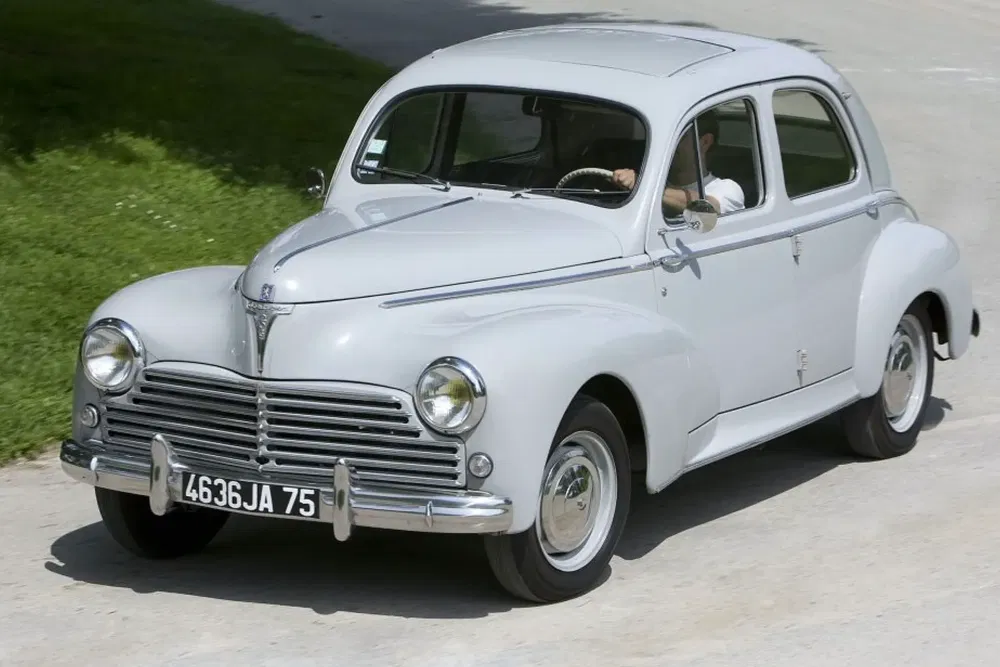 Peugeot 203 classique gris roulant sur une route rurale ensoleillée.