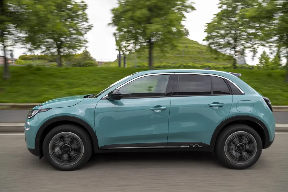 Lichtblauwe elektrische Kia EV5 SUV rijdend op groene weg met vervaagde bomen op achtergrond.