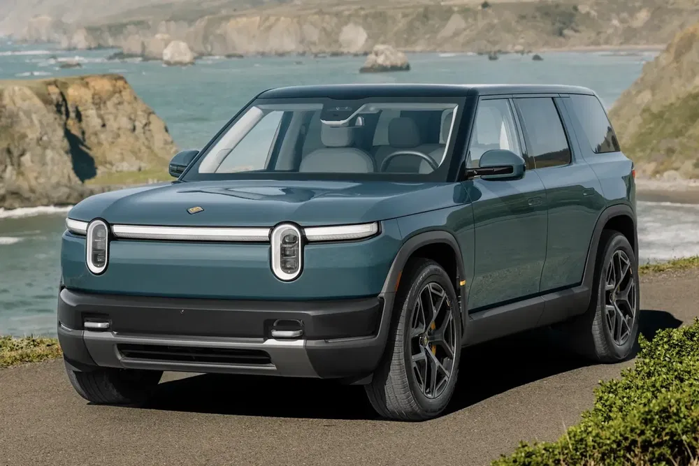 Rivian R1S SUV électrique sur route côtière avec vue sur l'océan et collines.