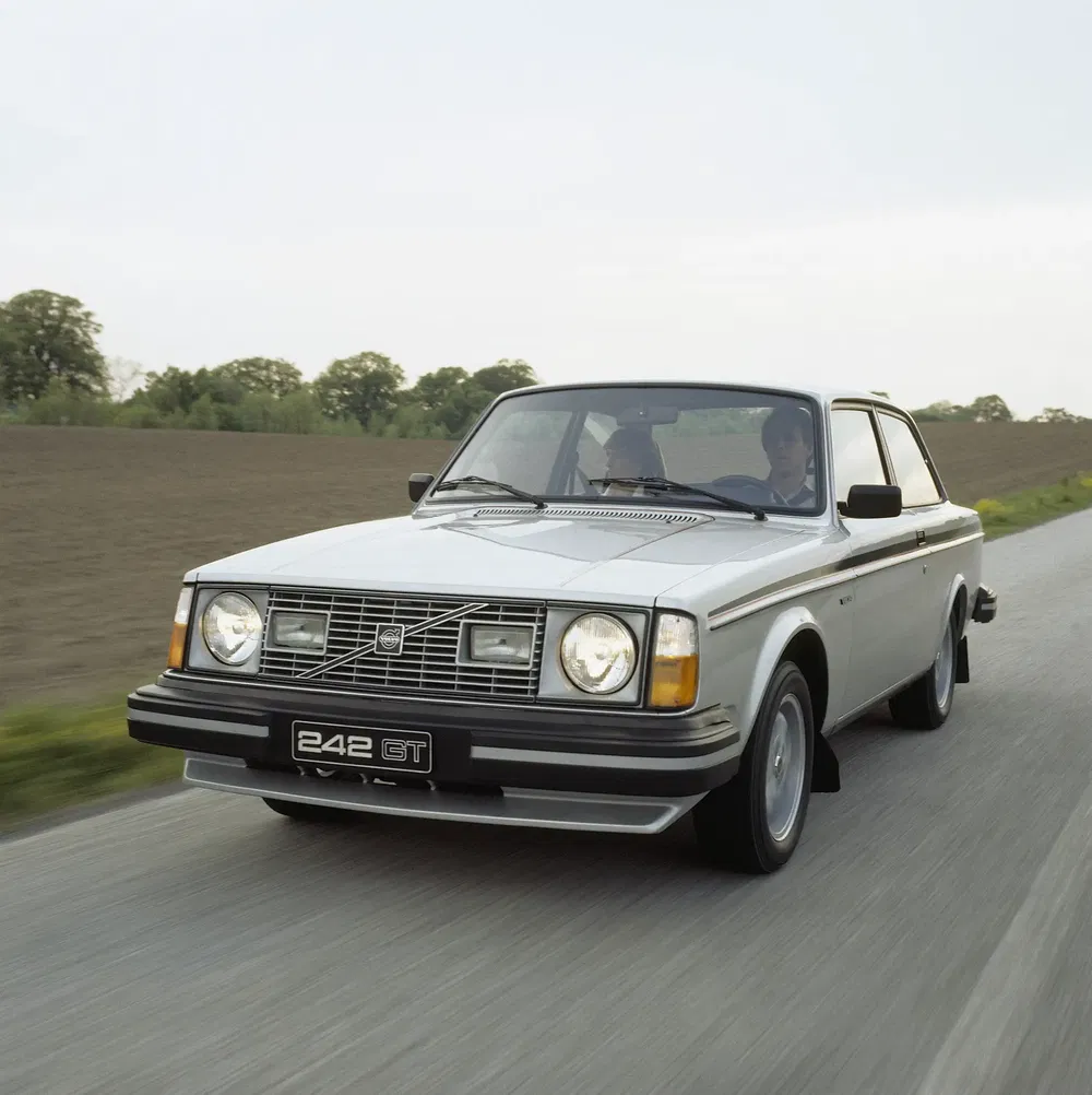 Volvo 242 GT argentée roulant sur une route de campagne bordée d'arbres, modèle rétro.