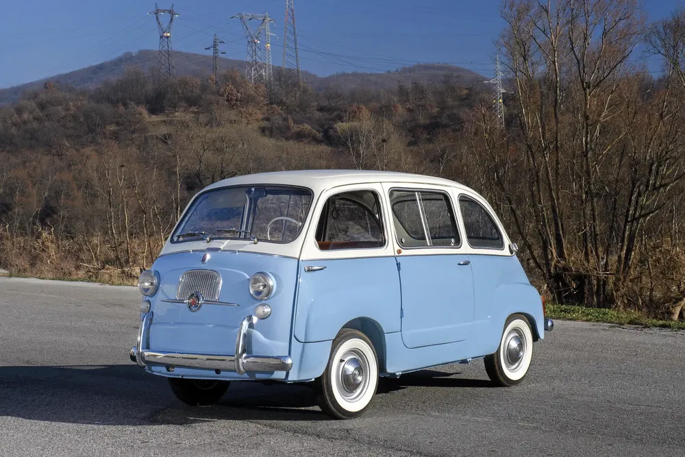 Fiat 600 Multipla vintage bleu ciel sur fond de colline arborée.
