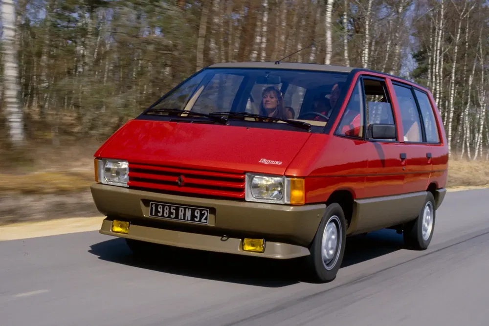 Renault Espace rouge de 1985 roulant sur une route bordée d'arbres, avant lumineux distinctif.