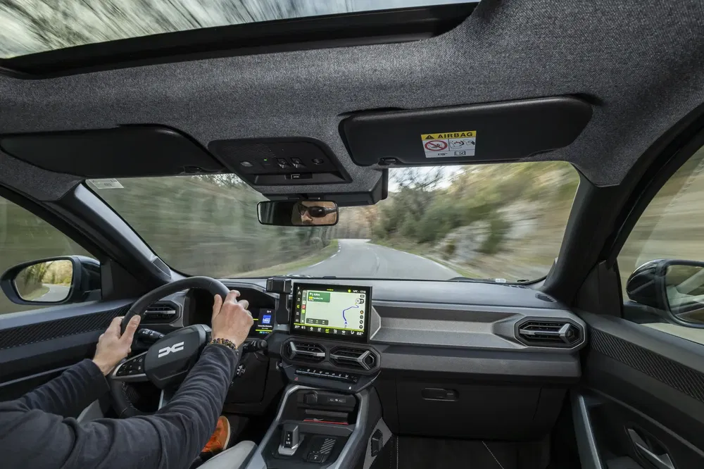 Bestuurder in een Dacia-auto met navigatiescherm op een kronkelige weg.