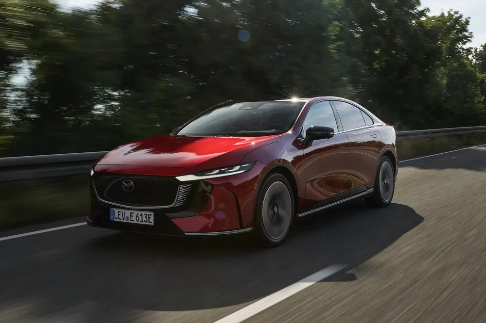 Rode Mazda-auto rijdt op een weg, omgeven door groen en onder een zonnige hemel.