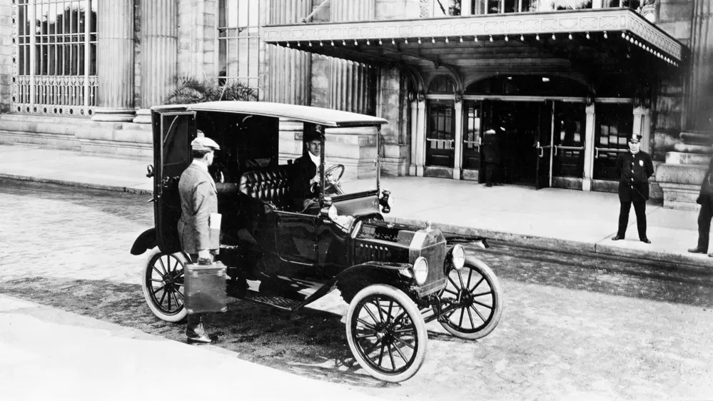 Voiture ancienne devant bâtiment historique avec chauffeur et passager, début du XXe siècle.