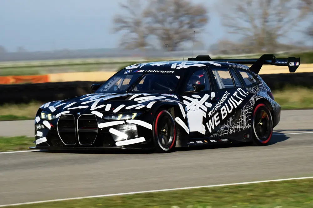 BMW auto met camouflage op racecircuit, voorzien van een grote zwarte achtervleugel.