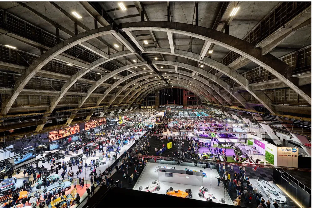 {"fr":"Vue aérienne d'un salon automobile rempli de stands et de visiteurs sous une structure voûtée.","nl":"Luchtfoto van een autobeurs met stands en bezoekers onder een gewelfde structuur."}