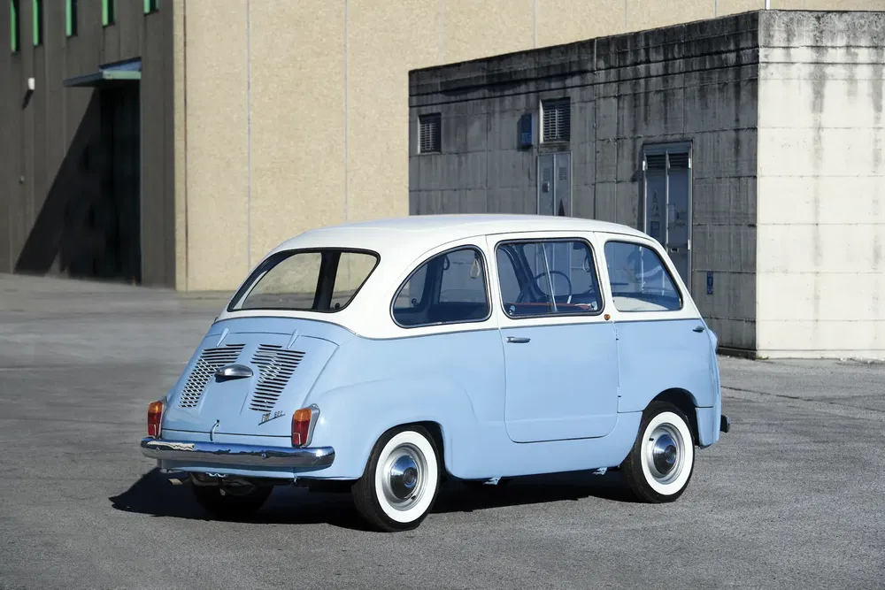 Fiat 600 Multipla bleu clair et blanc stationné devant un bâtiment industriel.