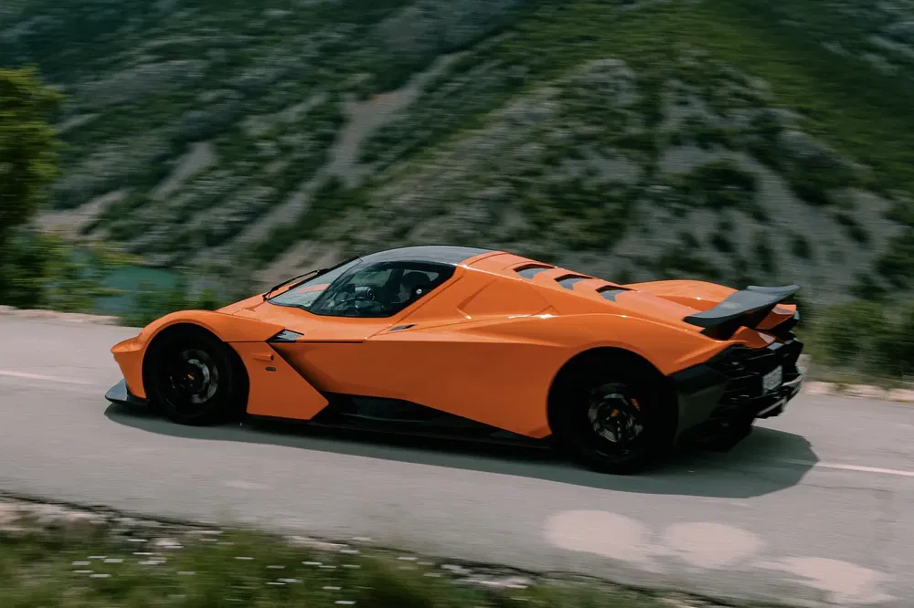 {"fr":"Supercar orange KTM X-Bow GT sur route montagneuse, paysage verdoyant en arrière-plan.","nl":"Oranje KTM X-Bow GT supercar op bergweg, groene heuvels op de achtergrond."}