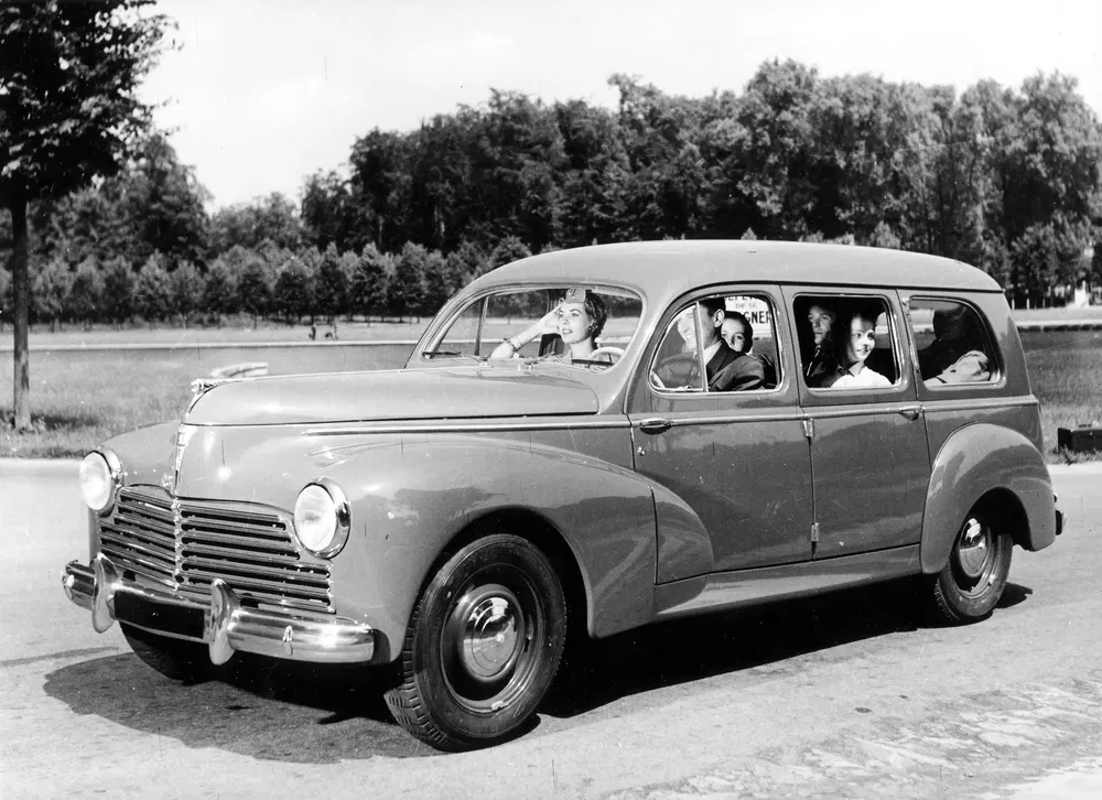 Peugeot 203 familiale des années 1950 avec quatre passagers sur une route arborée.
